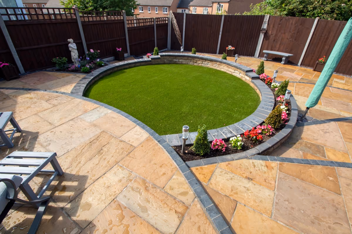 Three level camal natural stone patio with large charcoal rumbled block, steps built from charcoal kerbs, mellow York abbey walling containing a bedding area and artificial grass. -angle 1
