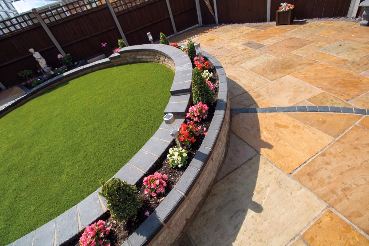 Three level camal natural stone patio with large charcoal rumbled block, steps built from charcoal kerbs, mellow York abbey walling containing a bedding area and artificial grass. -angle 6