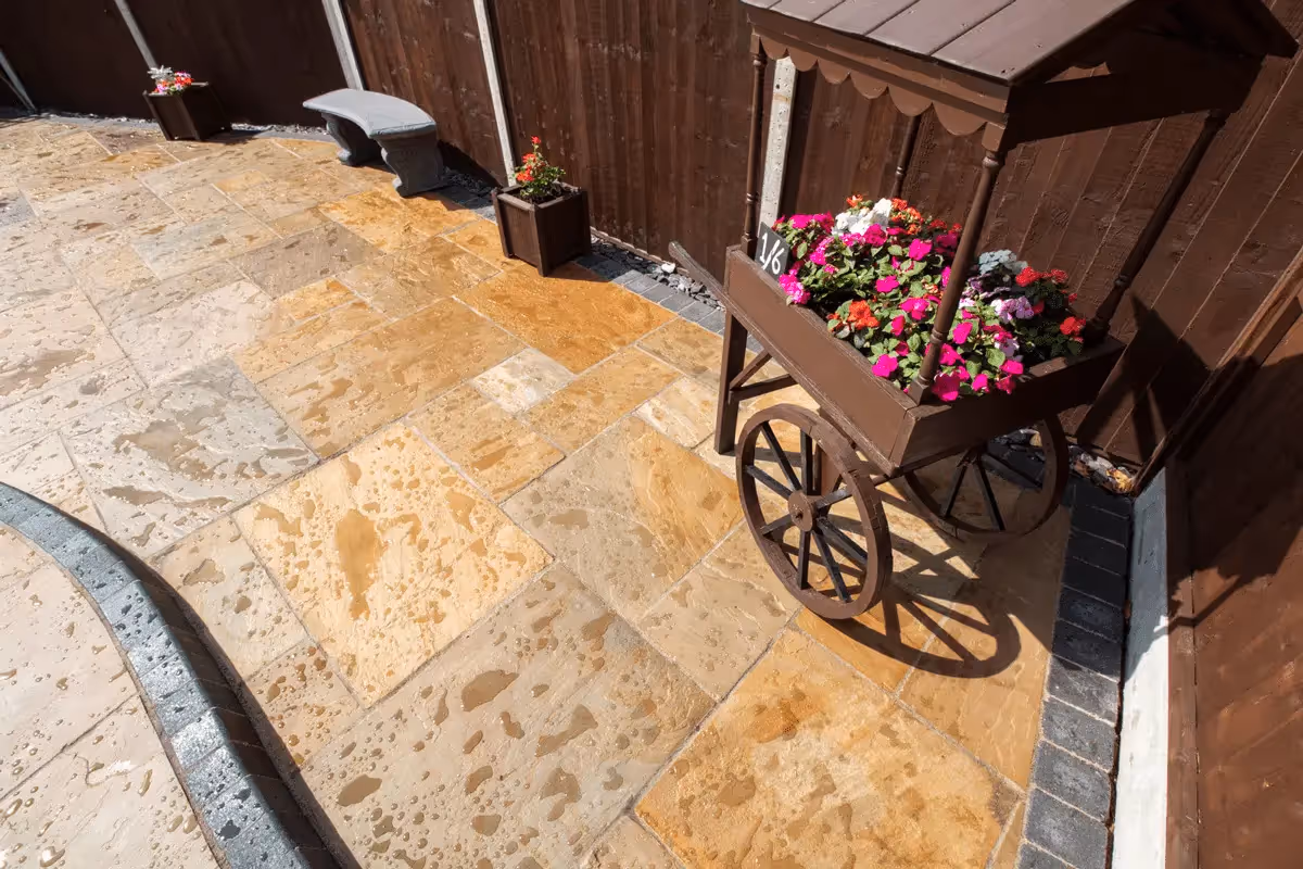 Three level camal natural stone patio with large charcoal rumbled block, steps built from charcoal kerbs, mellow York abbey walling containing a bedding area and artificial grass. -angle 5