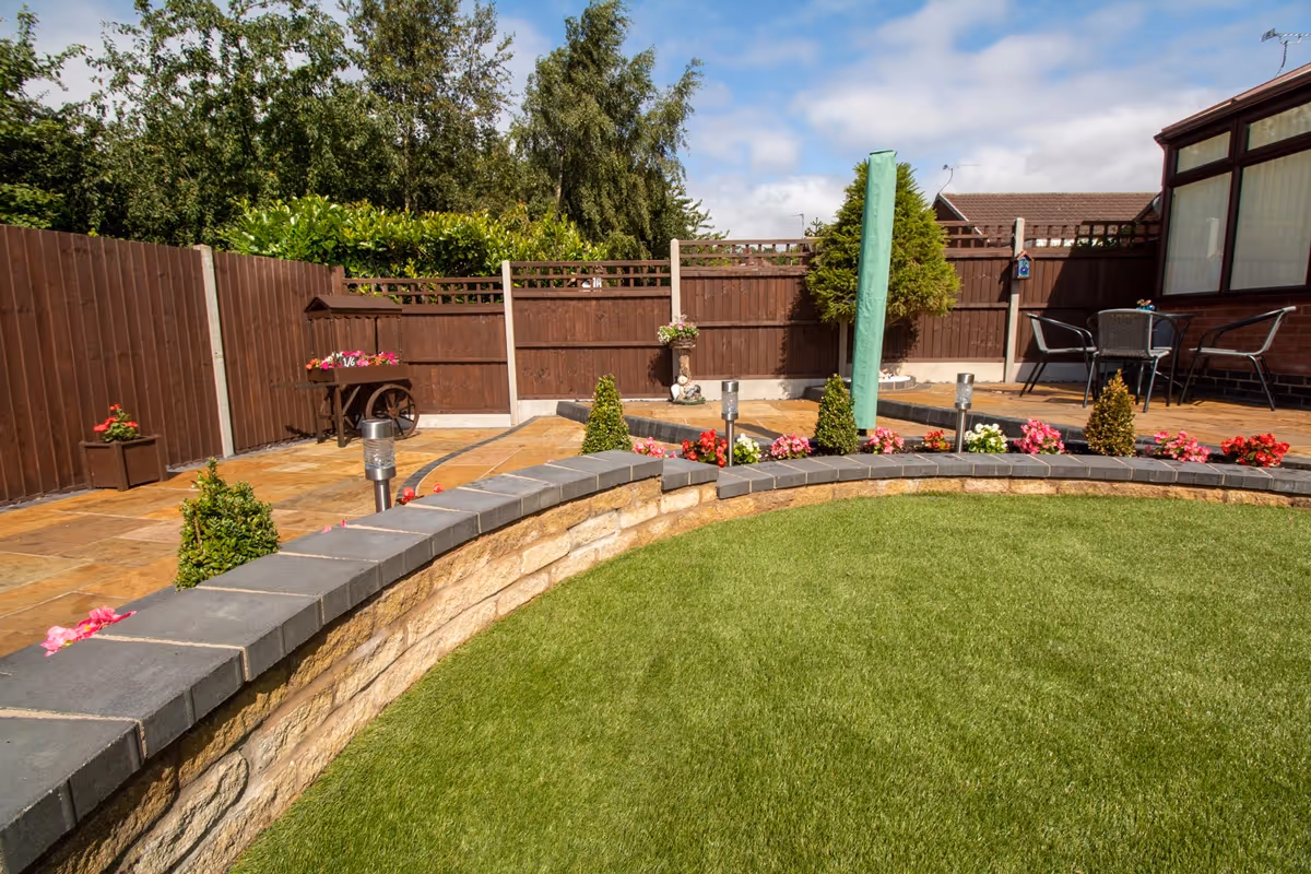 Three level camal natural stone patio with large charcoal rumbled block, steps built from charcoal kerbs, mellow York abbey walling containing a bedding area and artificial grass. -angle 4