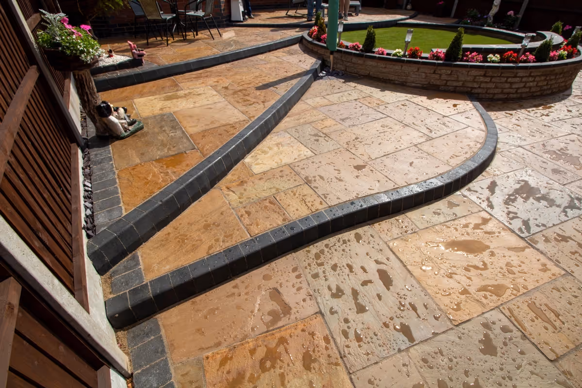 Three level camal natural stone patio with large charcoal rumbled block, steps built from charcoal kerbs, mellow York abbey walling containing a bedding area and artificial grass. -angle 7