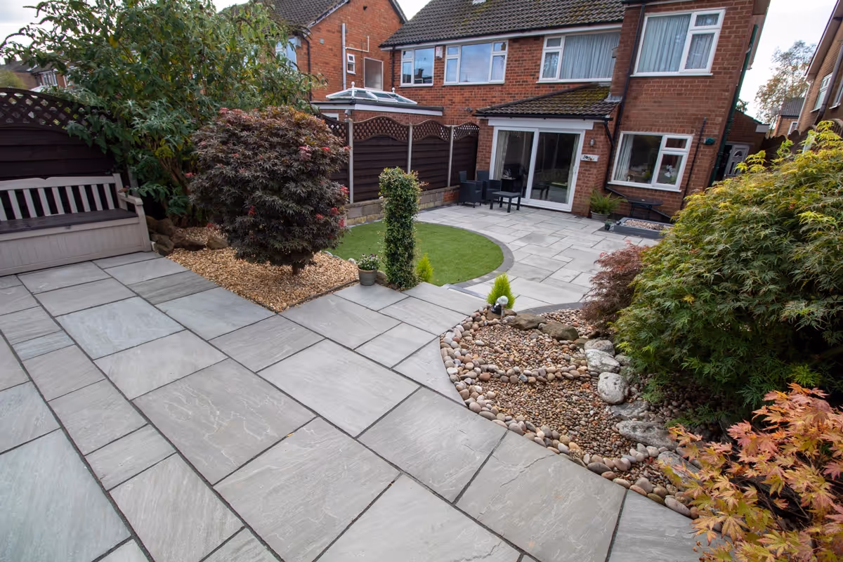 Mixed size grey sandstone patio with a large charcoal rumbled block border, artificial grass also edged with charcoal blocks. 