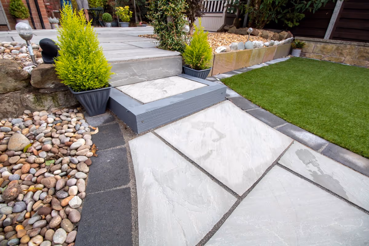 Mixed size grey sandstone patio with a large charcoal rumbled block border, artificial grass also edged with charcoal blocks.  - angle 1