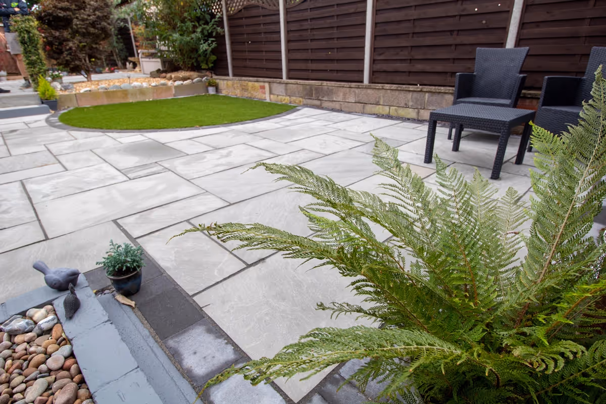 Mixed size grey sandstone patio with a large charcoal rumbled block border, artificial grass also edged with charcoal blocks.  - angle 6