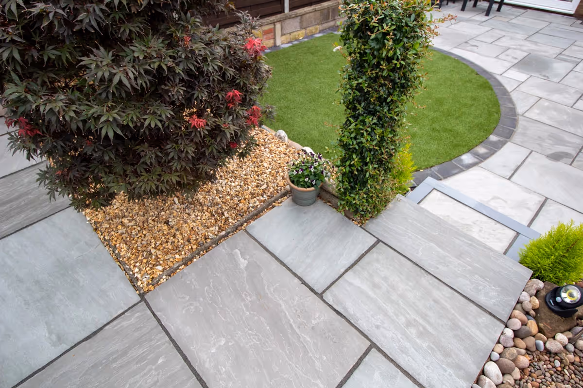 Mixed size grey sandstone patio with a large charcoal rumbled block border, artificial grass also edged with charcoal blocks.  - angle 5
