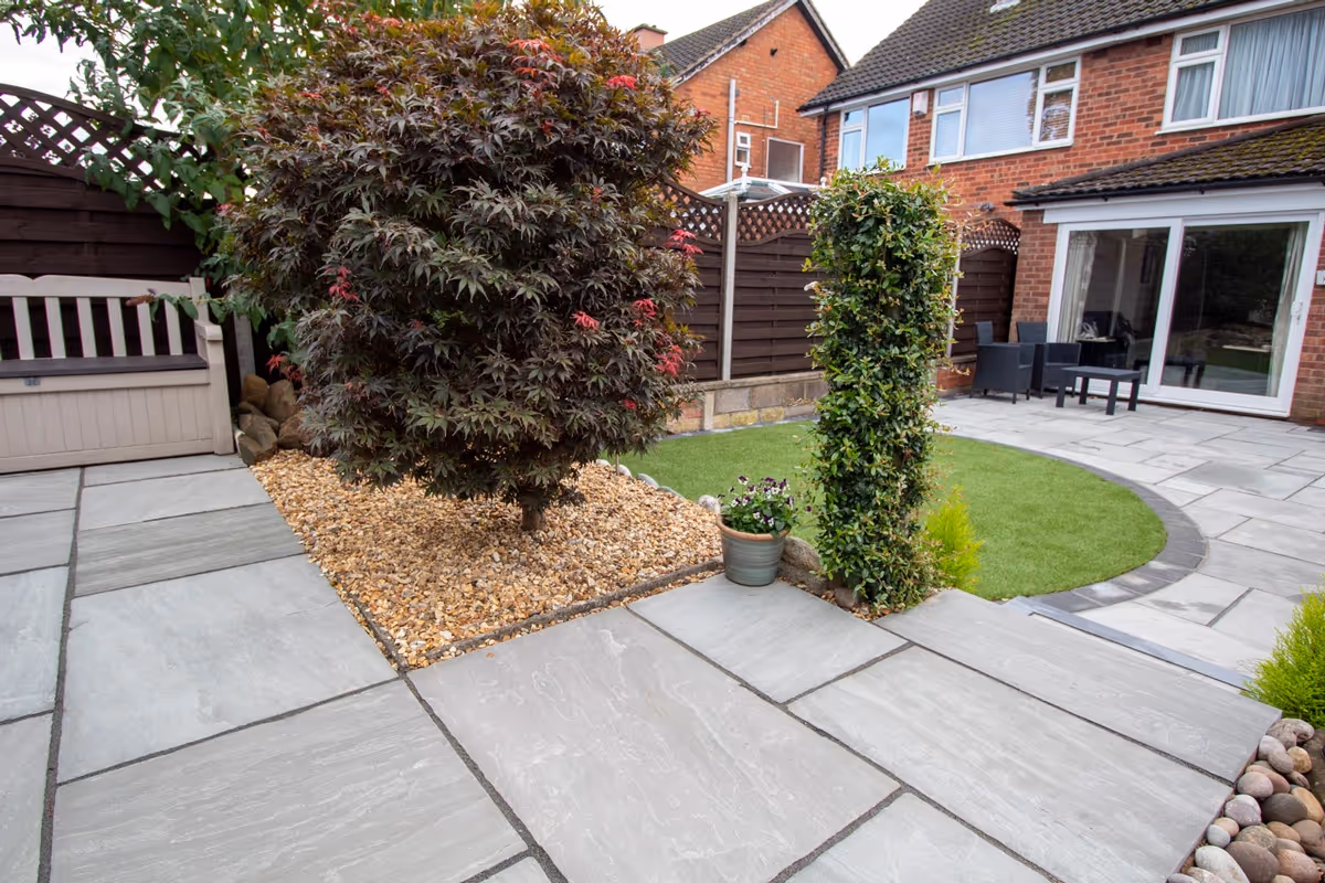 Mixed size grey sandstone patio with a large charcoal rumbled block border, artificial grass also edged with charcoal blocks.  - angle 7