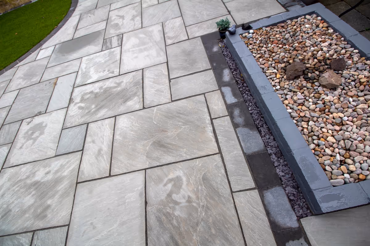 Mixed size grey sandstone patio with a large charcoal rumbled block border, artificial grass also edged with charcoal blocks.  - angle 3