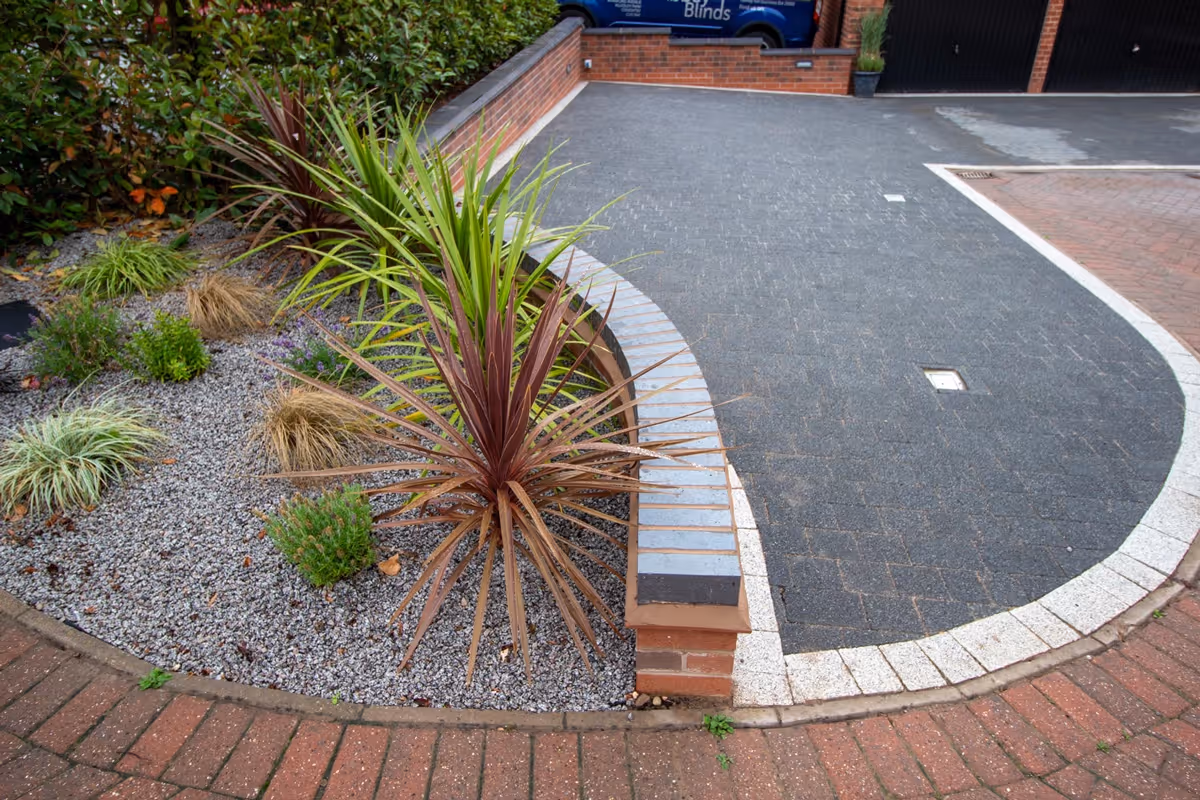 Resin-topped regular block paving driveway with white block border and security posts for a caravan. Retaining wall with tile crease and blue engineering header course - angle 1