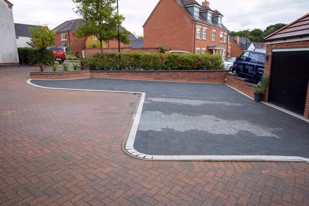 Resin-topped regular block paving driveway with white block border and security posts for a caravan. Retaining wall with tile crease and blue engineering header course - angle 3
