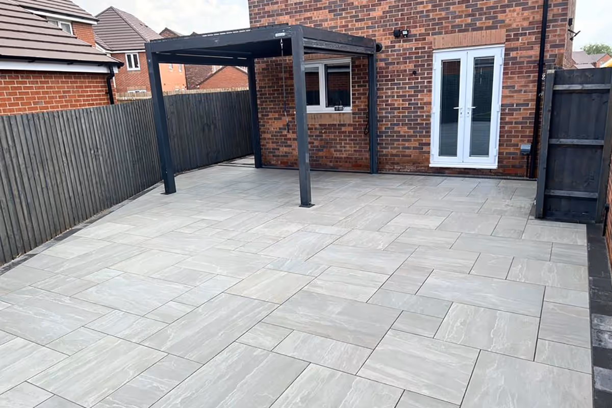 Large Kandla mixed-size porcelain patio with charcoal pencil edge paving. Charcoal KL kerb set. ACO drains. 4x3m Pergolux Aluminium Pergola in Anthracite. Pet-friendly artificial turf.
