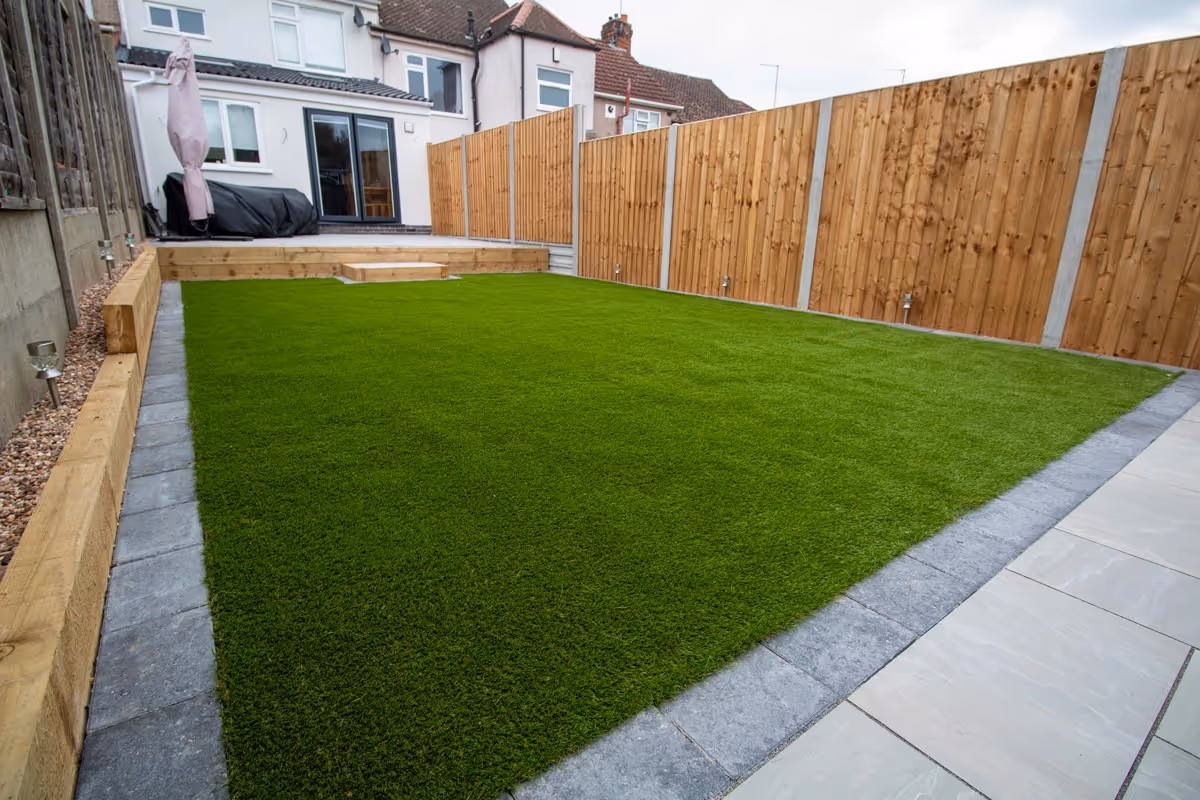 Kandla mixed-size porcelain slabs with charcoal pencil edge paving. Fence with concrete posts and gravel boards. Sleeper retainer. Artificial turf.