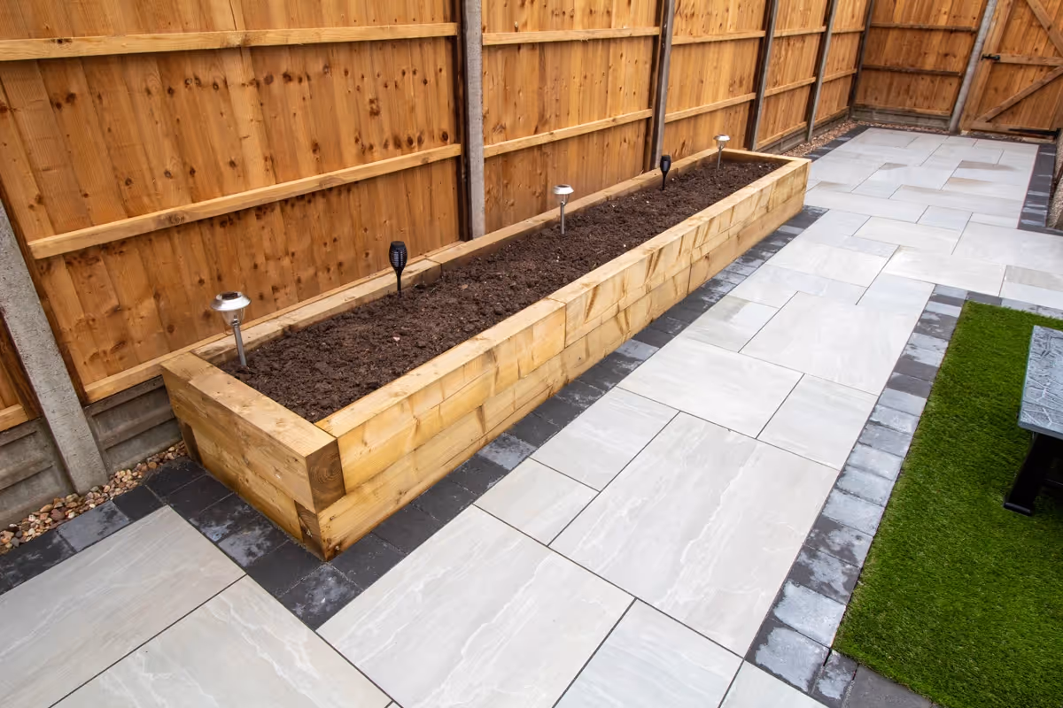 Kandla mixed-size porcelain patio with Charcoal pencil edge paving. Raised sleeper planters. Artificial Turf. New fence and gate - angle 1