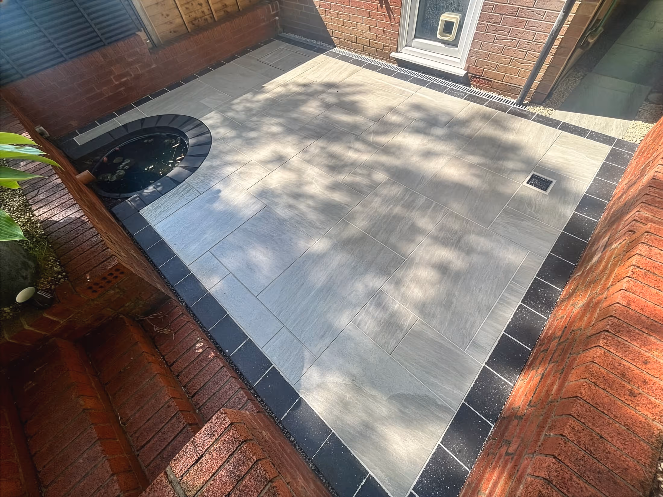 Small patio with light gray tiles bordered by dark tiles, a round pond with lilies, and red brick walls with a cat flap door.