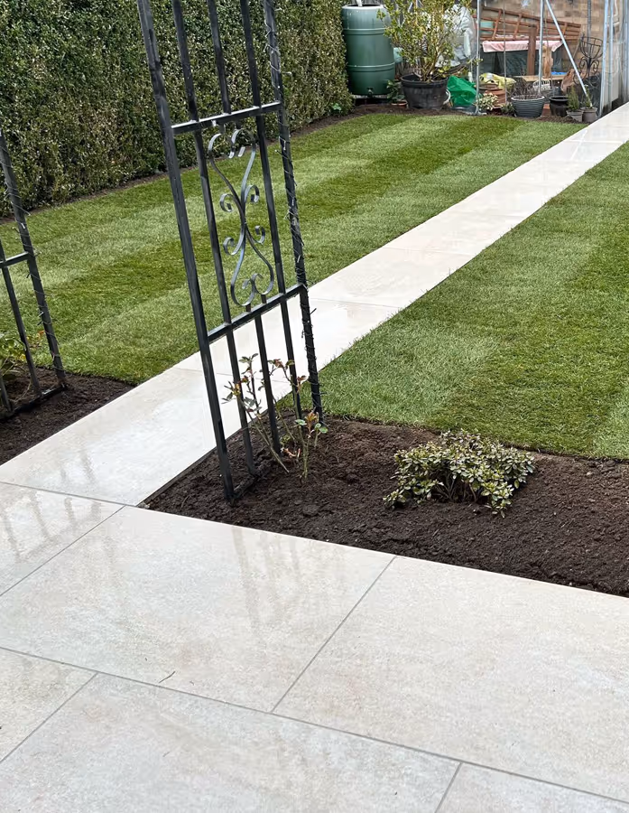 Garden with trimmed grass, a paved walkway, a metal garden arch, and a few small plants in soil beds.