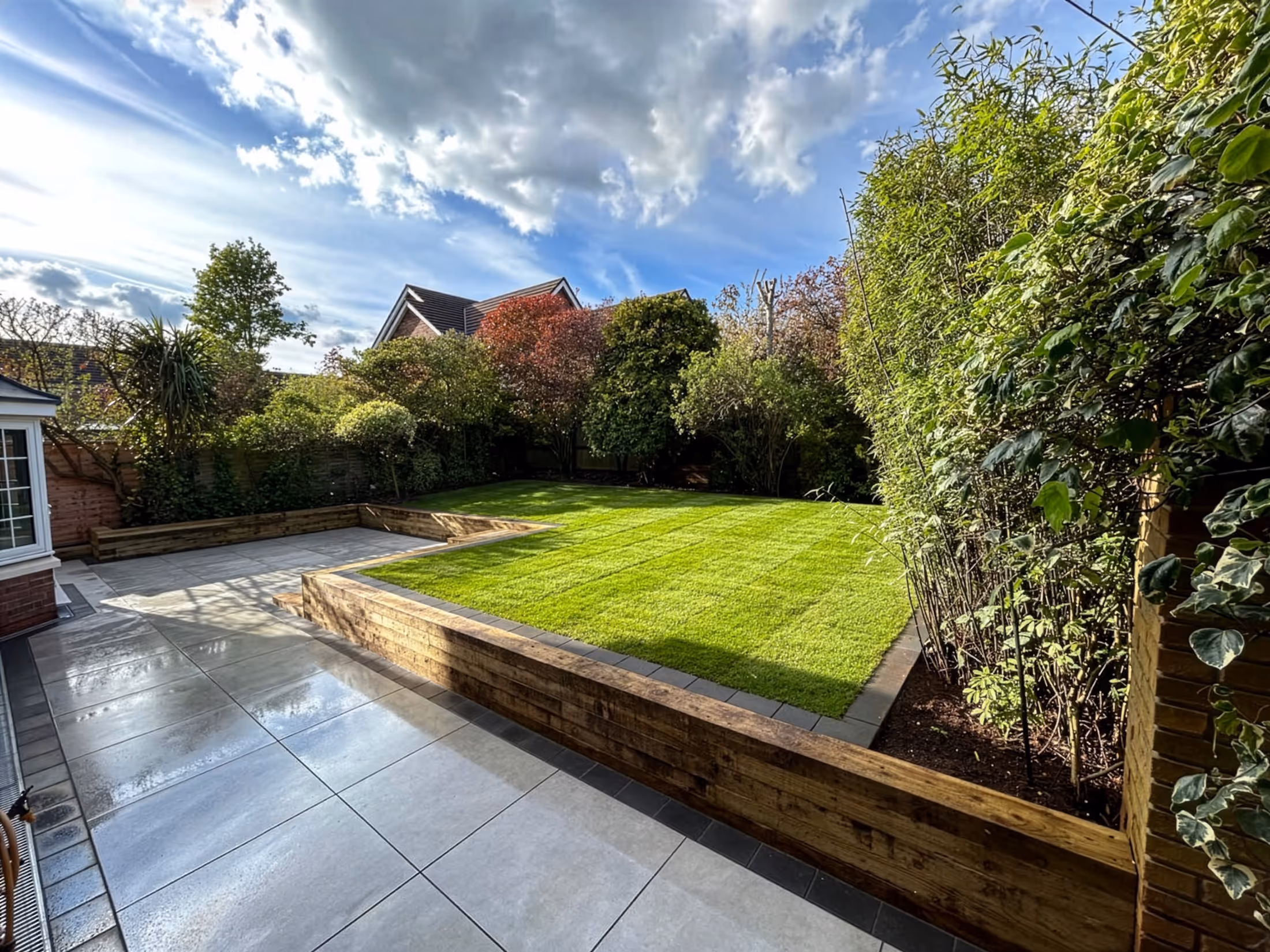 Neatly landscaped backyard with fresh green lawn, wooden retaining walls, stone tiled patio, and surrounding trees under a partly cloudy sky.