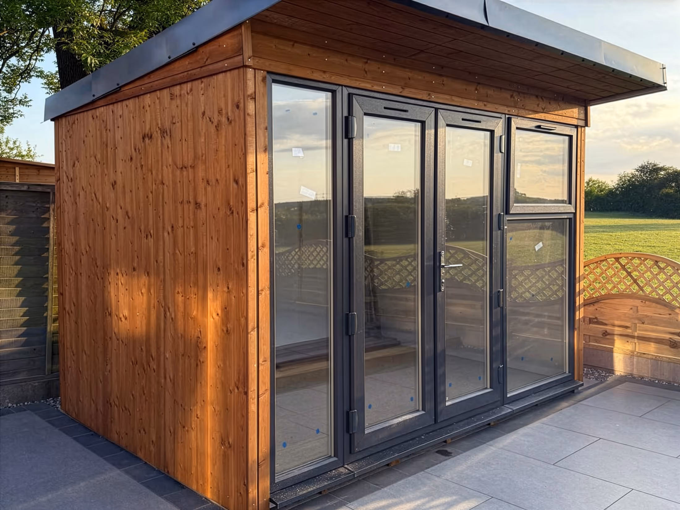 Modern wooden garden room with large dark-framed glass doors and windows on a stone patio.