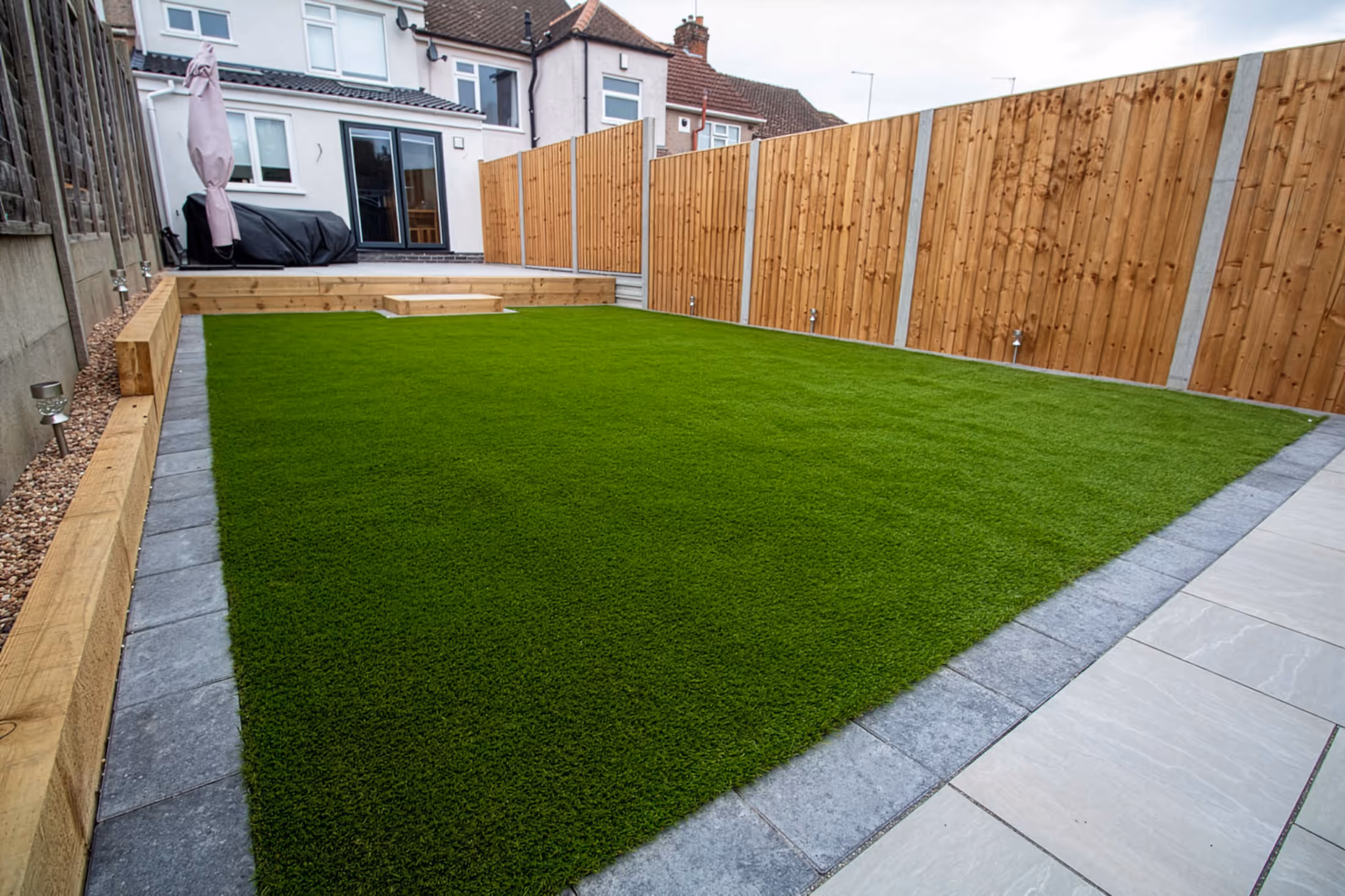 Well-maintained backyard with artificial green grass bordered by gray stone tiles and wooden fences.