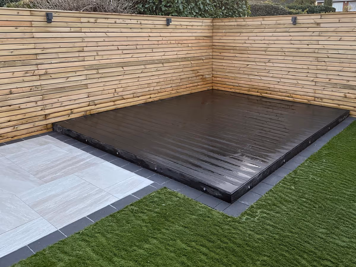 Corner backyard with light gray stone tiles, artificial green grass, and a raised glossy black wooden platform against a wooden slat fence.