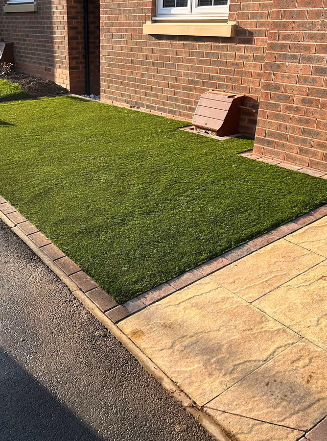Small front yard with a rectangular patch of artificial grass bordered by brick edging, next to a paved stone walkway and a brick house wall.
