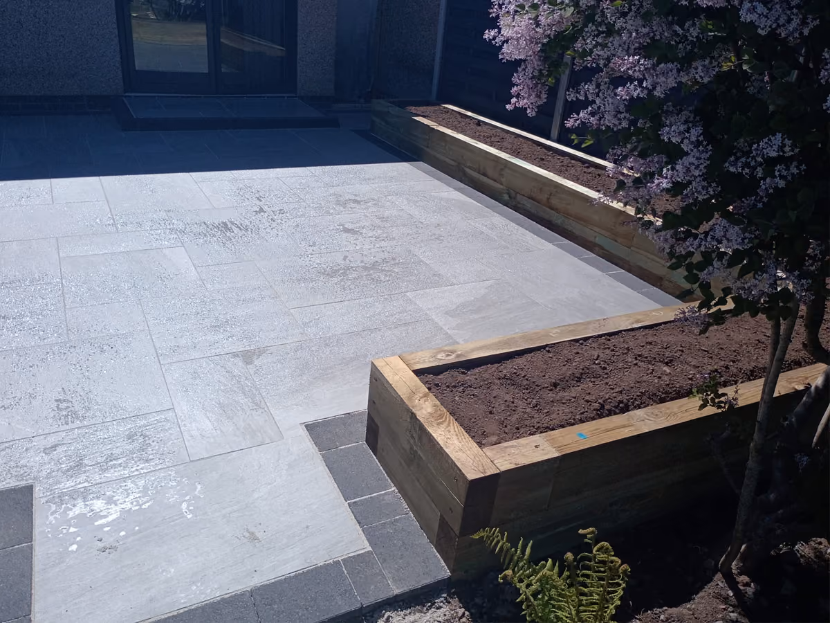 Outdoor tiled patio with raised wooden garden beds, soil, and a flowering tree in sunlight.