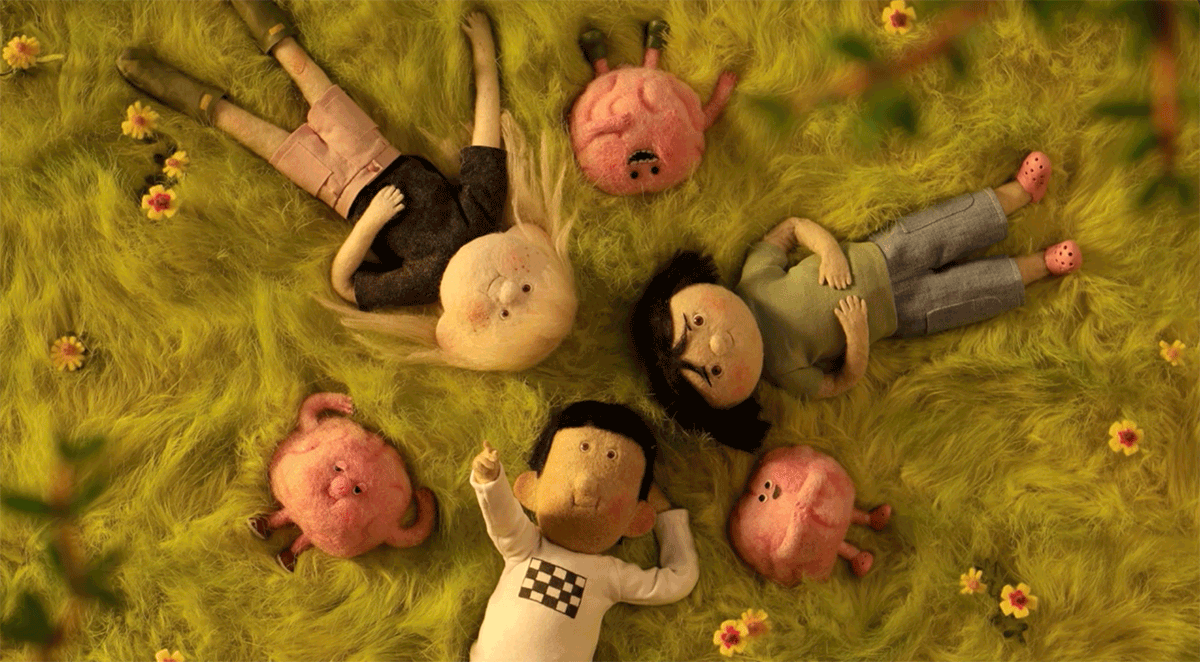 Three felt animated children and three pink felt brains lying on a fluffy green grass with small yellow flowers looking up at the sky.