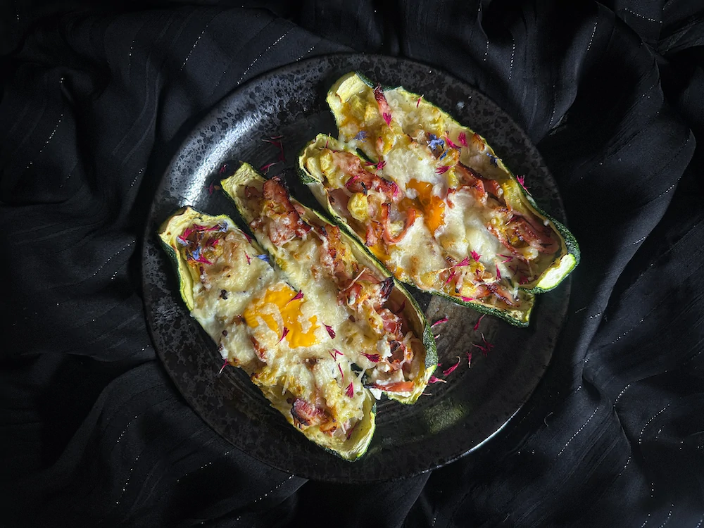 Les courgettes sont farcies de lardons, oeufs et fromage râpé et elles sont placées sur une assiette plate rose. 