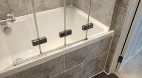 Completed bathroom with a white bathtub, grey tiled panel and glass shower screen