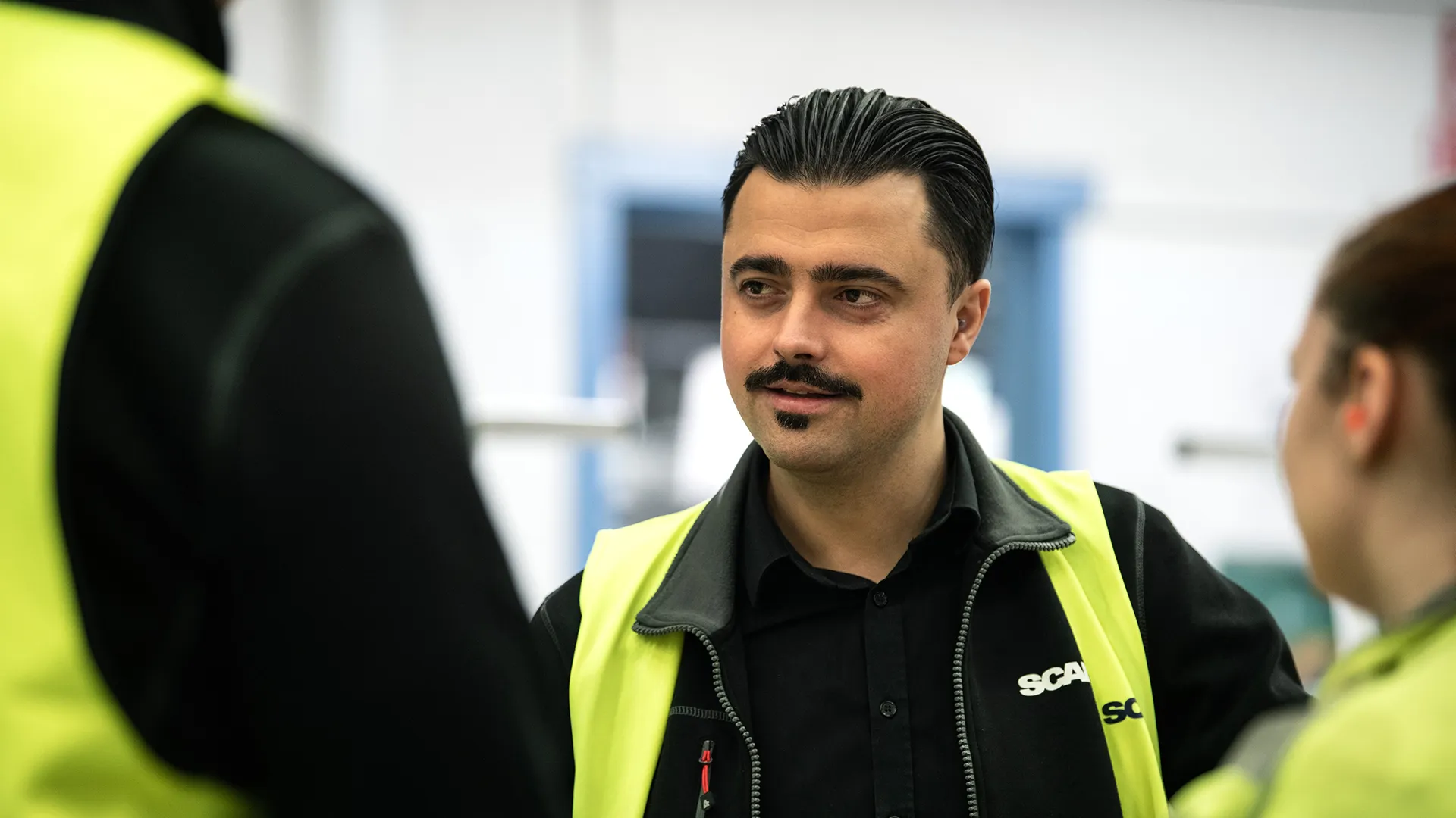 Man med mustasch och mörka och gula arbetskläder med Scanias logga på bröstet.