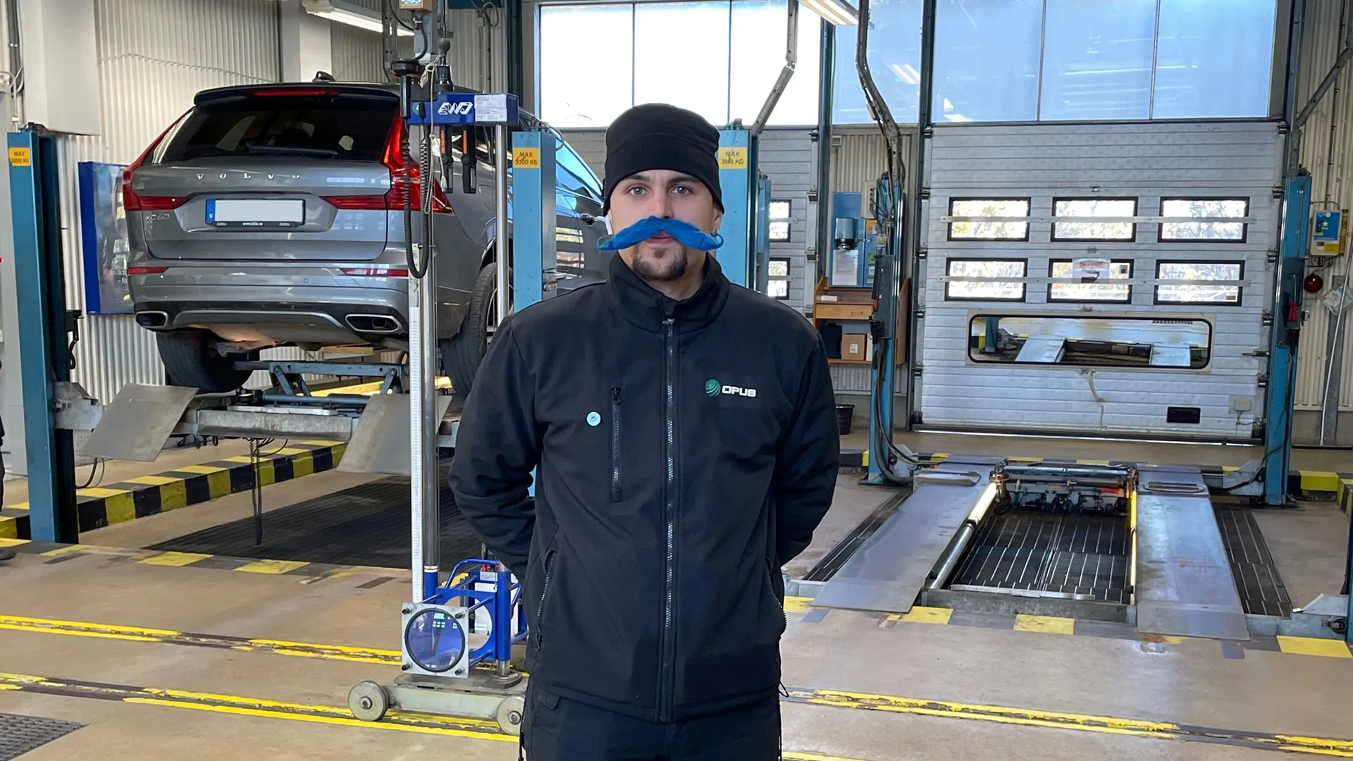 Ung man med stor blå lösmustasch iklädd mörka arbetskläder står i en verkstadshall för bilbesiktning.