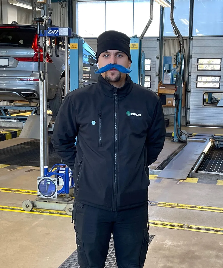 Ung man med stor blå lösmustasch iklädd mörka arbetskläder står i en verkstadshall för bilbesiktning.