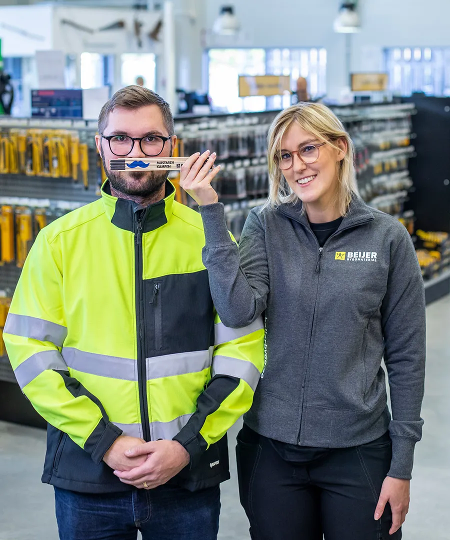 En man och en kvinna står inne i en bygghandel, kvinnan håller upp en tumstock med mustasch.