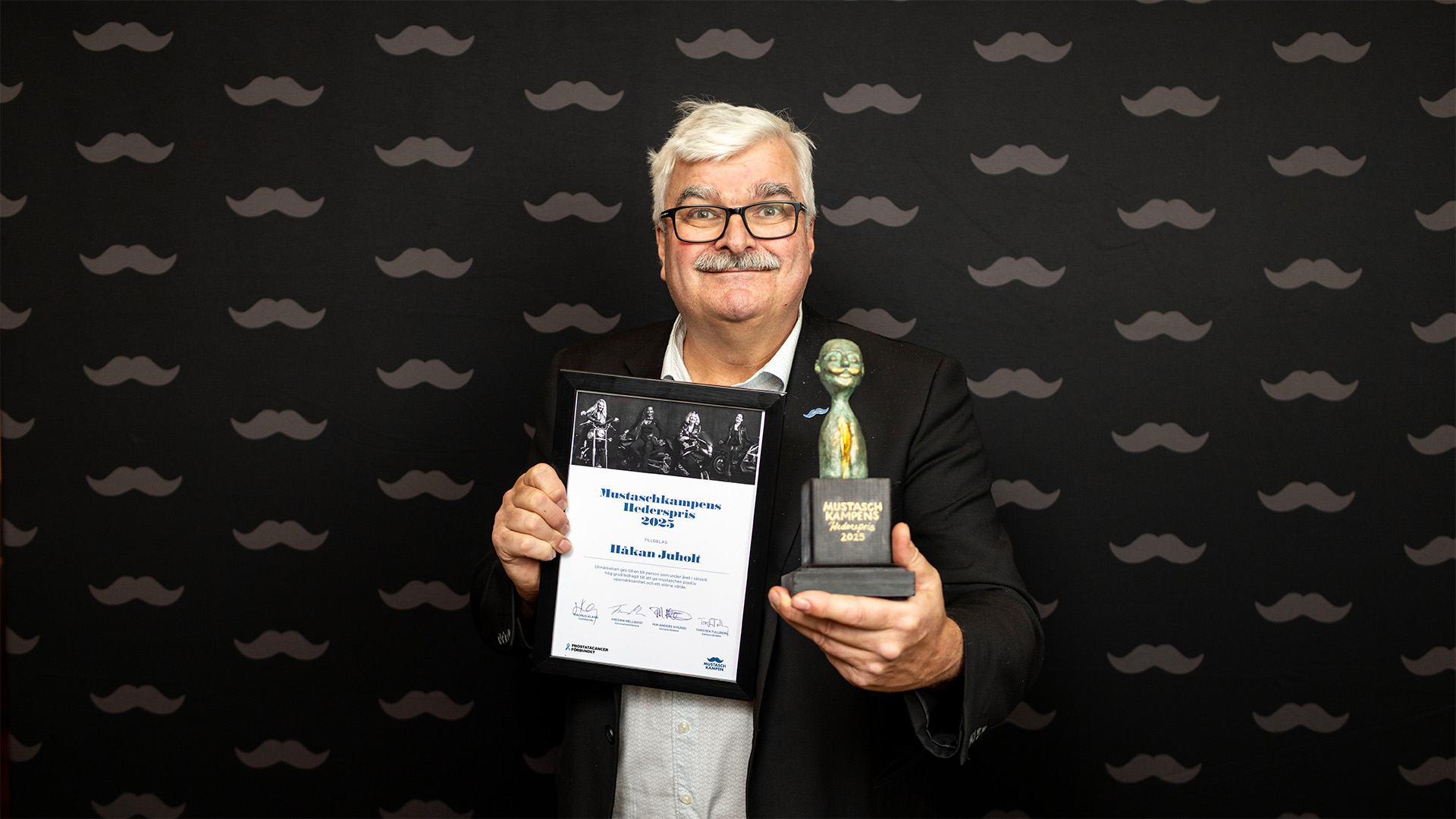 Man med mustasch som håller ett diplom och en skulptur framför sig