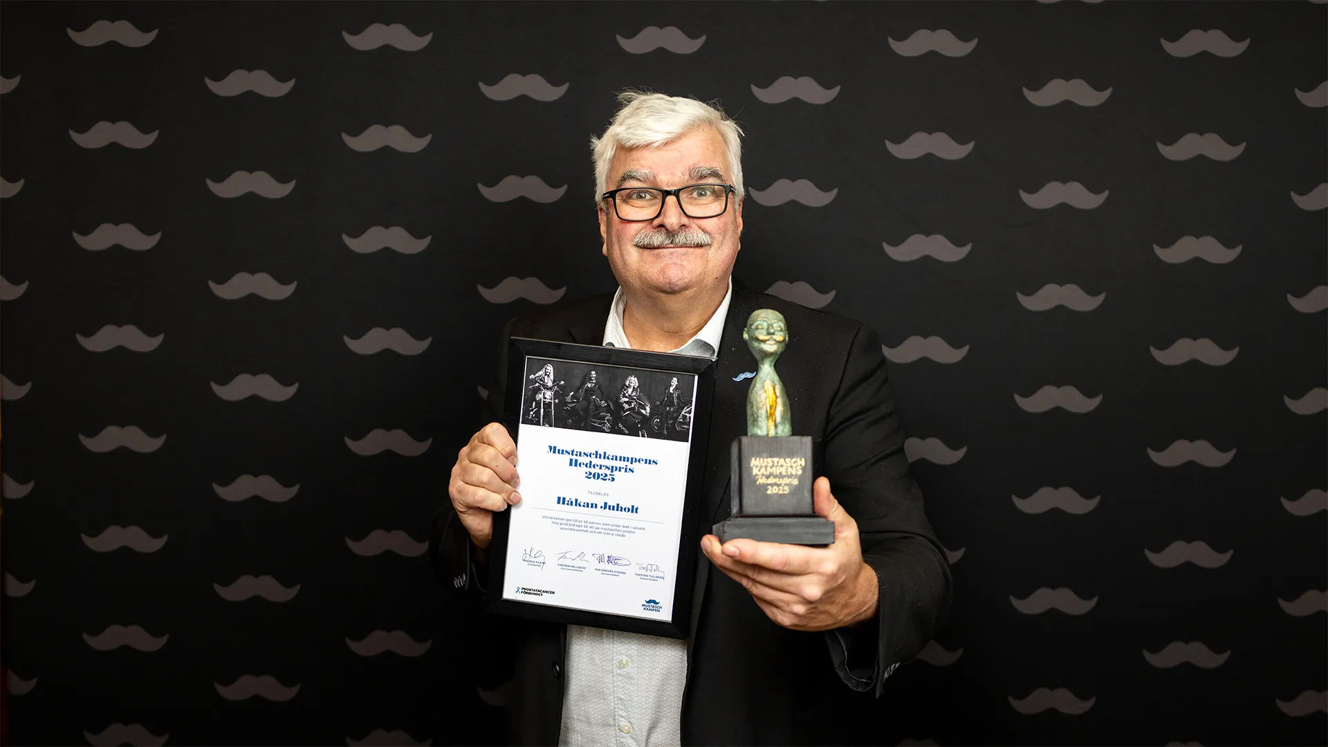 Man med mustasch som håller ett diplom och en skulptur framför sig