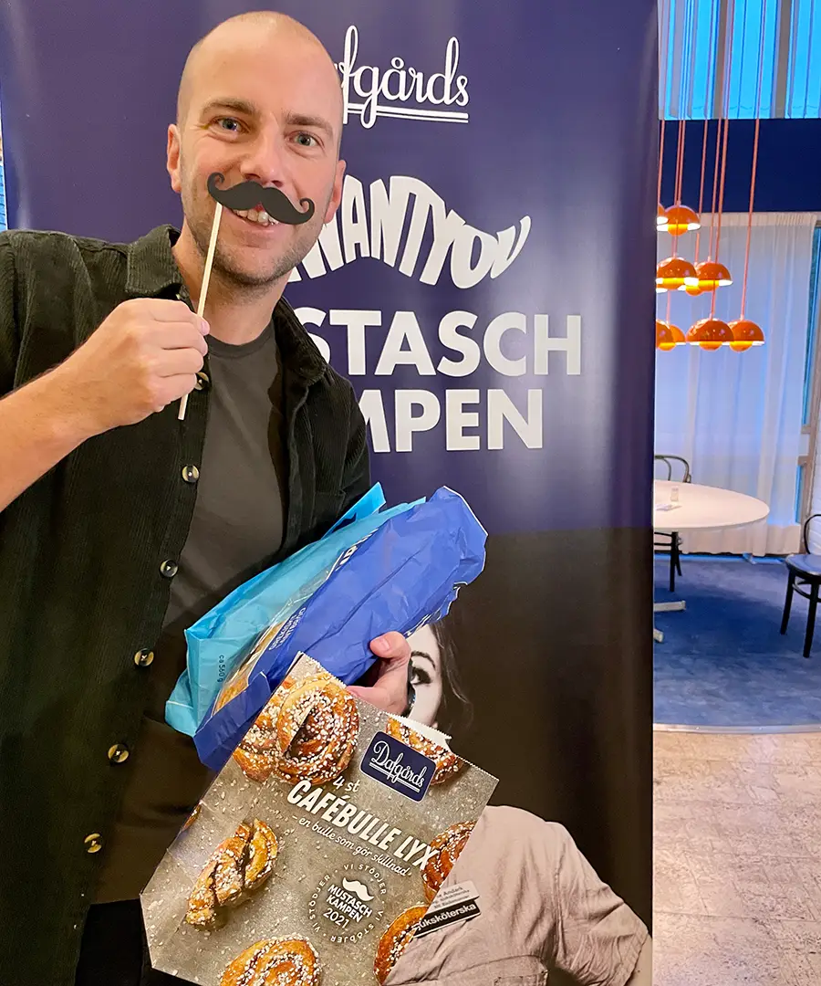 Man håller upp en lösmustasch och påsar med Dagårds mustaschprodukter.