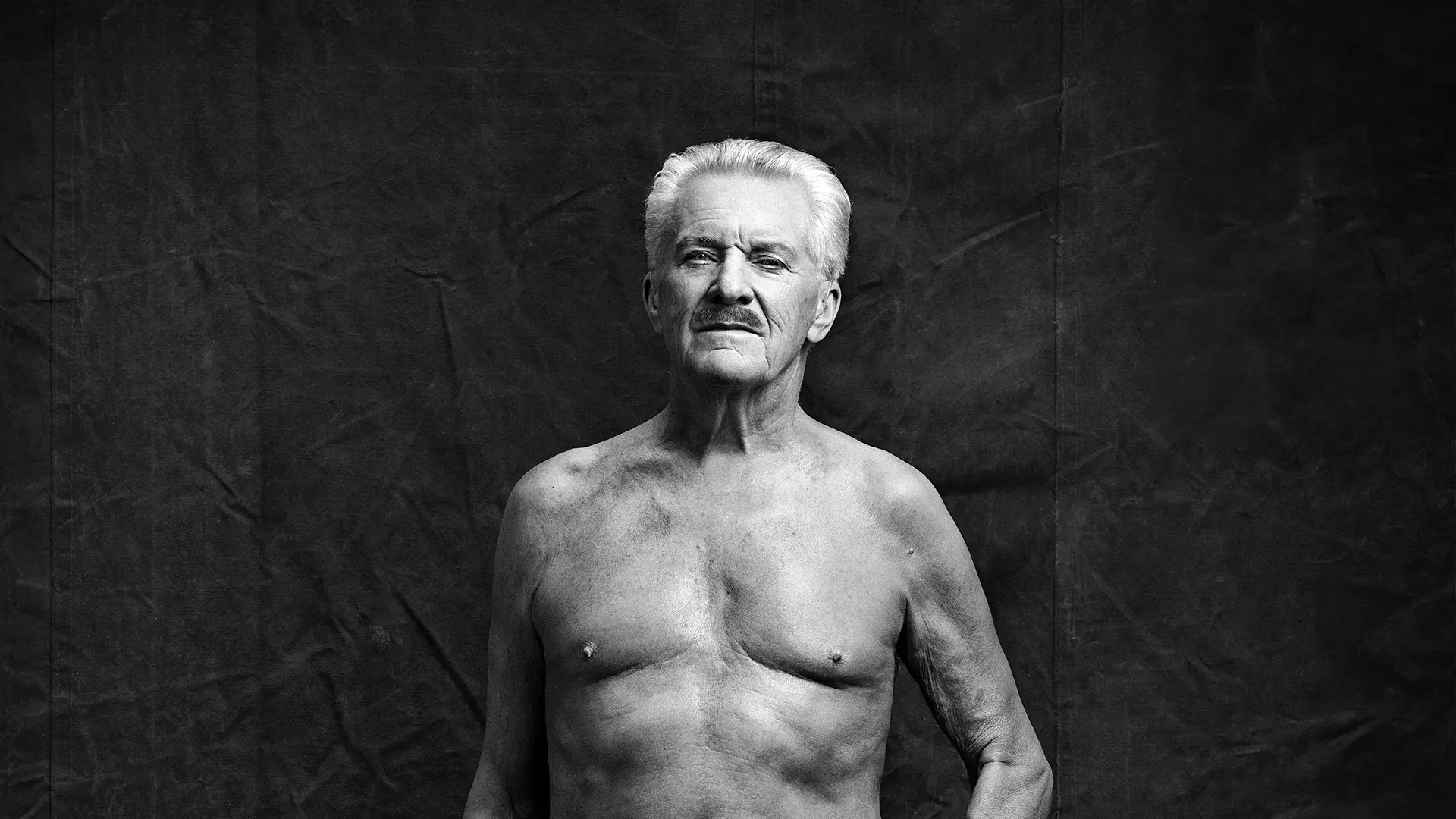 Porträttbild på äldre man i mustasch som är avklädd på överkroppen