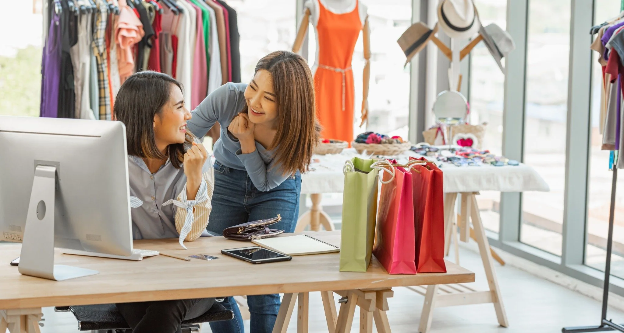 Two women smiling and talking at a desk with shopping bags in a clothing store setting.
