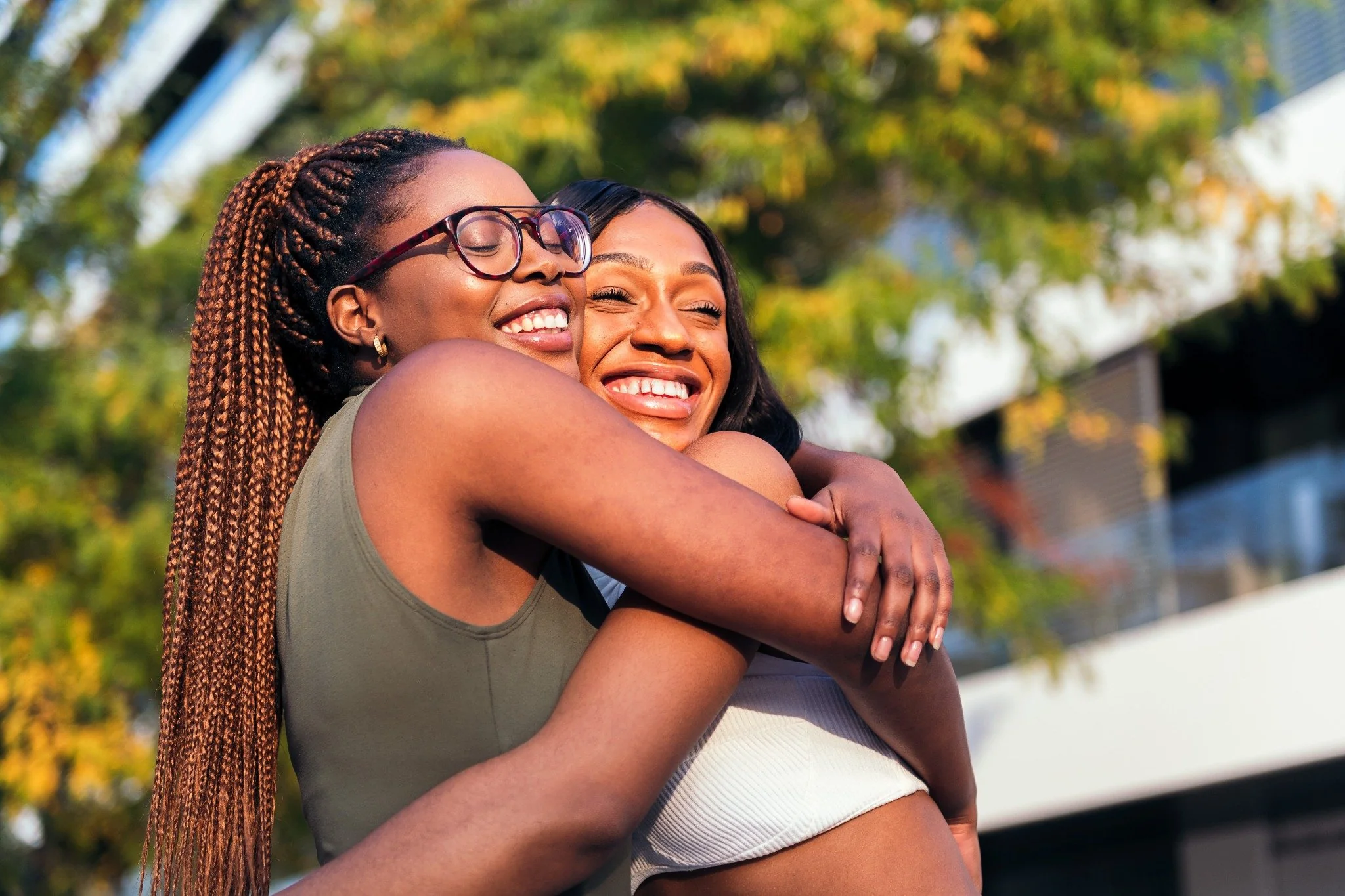 Two women smiling warmly while hugging outdoors with greenery and buildings in the background.