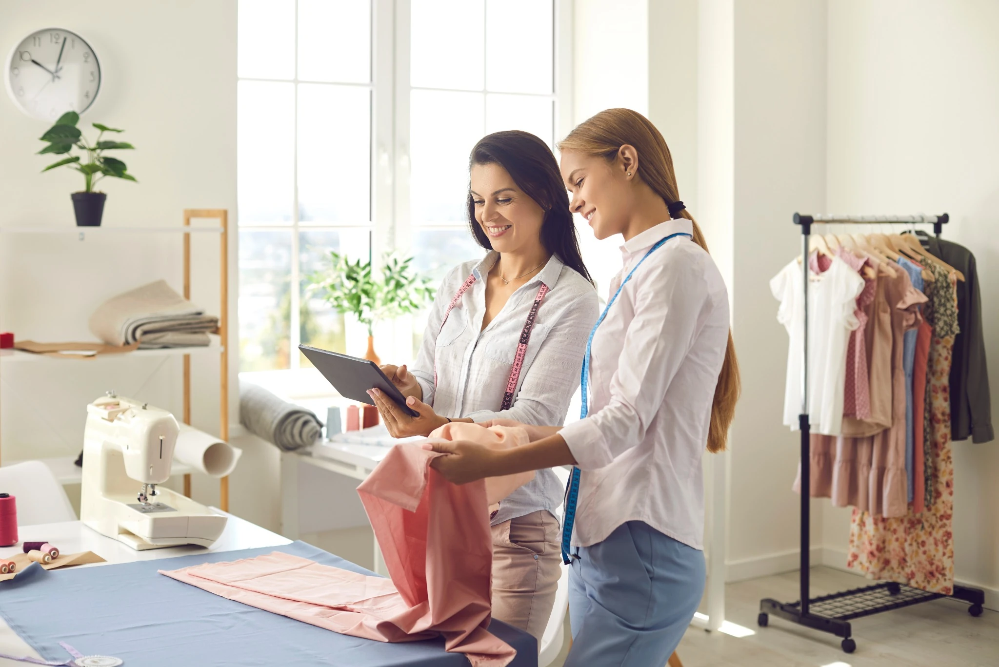 Two women with measuring tapes smiling, working with fabric and tablet in a bright sewing studio.