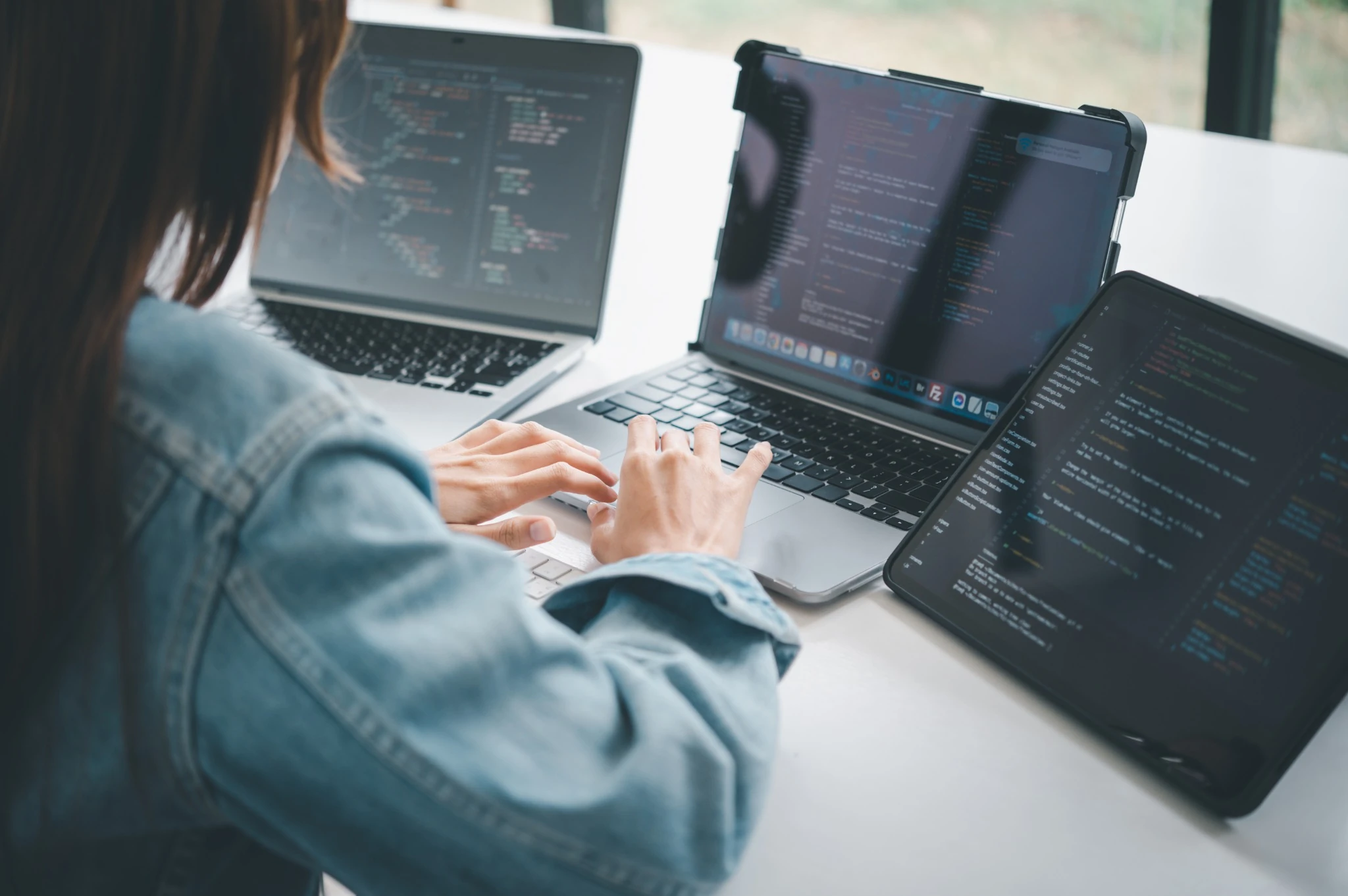 Person typing code on laptop with coding visible on two other devices on a white desk.