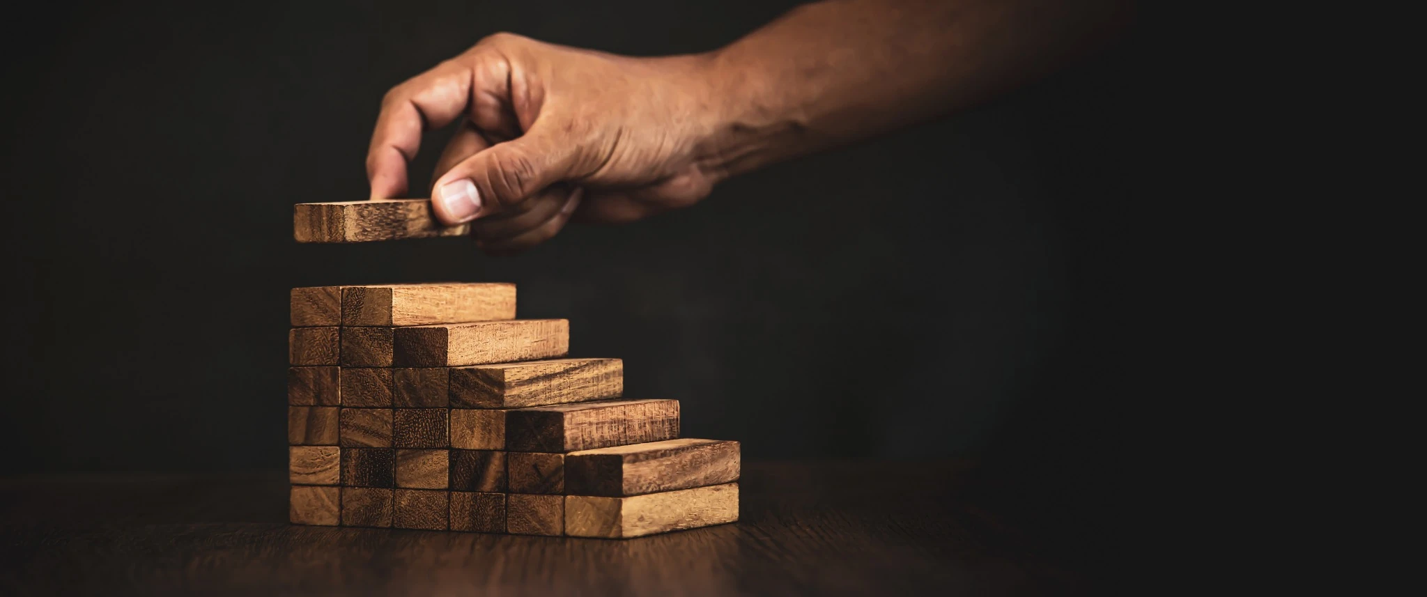 Hand placing a wooden block on top of a staircase made of wooden blocks on a dark background.
