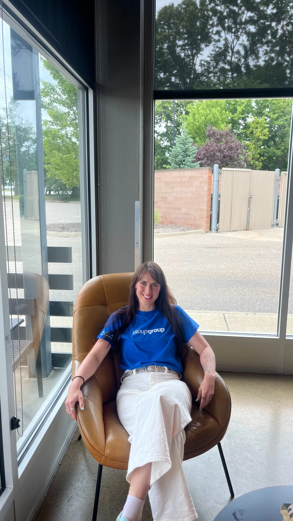 Woman in blue Shoup Group shirt sitting relaxed in a brown chair by a large window with street view.