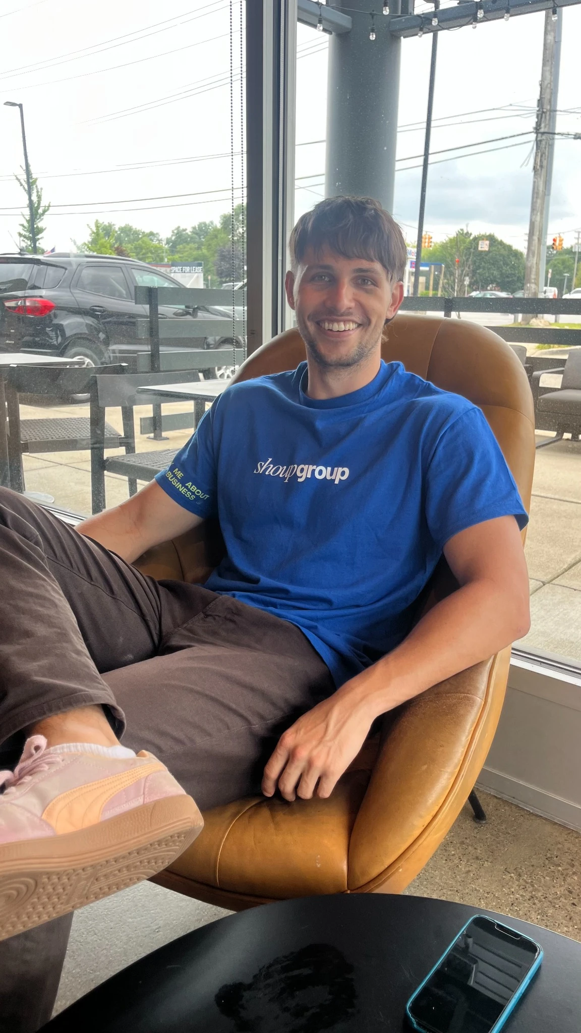 Smiling man in blue shirt sitting in a brown chair next to a window with a phone on a black table.