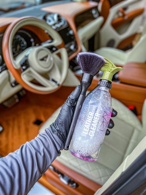 Gloved hand holding a bottle of O2M leather cleaner and a soft brush inside a car with tan and cream leather seats and steering wheel.