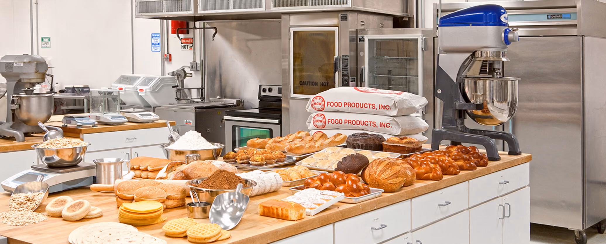 Commercial formula innovation bakery kitchen countertop with baked goods, flour, ingredients, and industrial mixers.