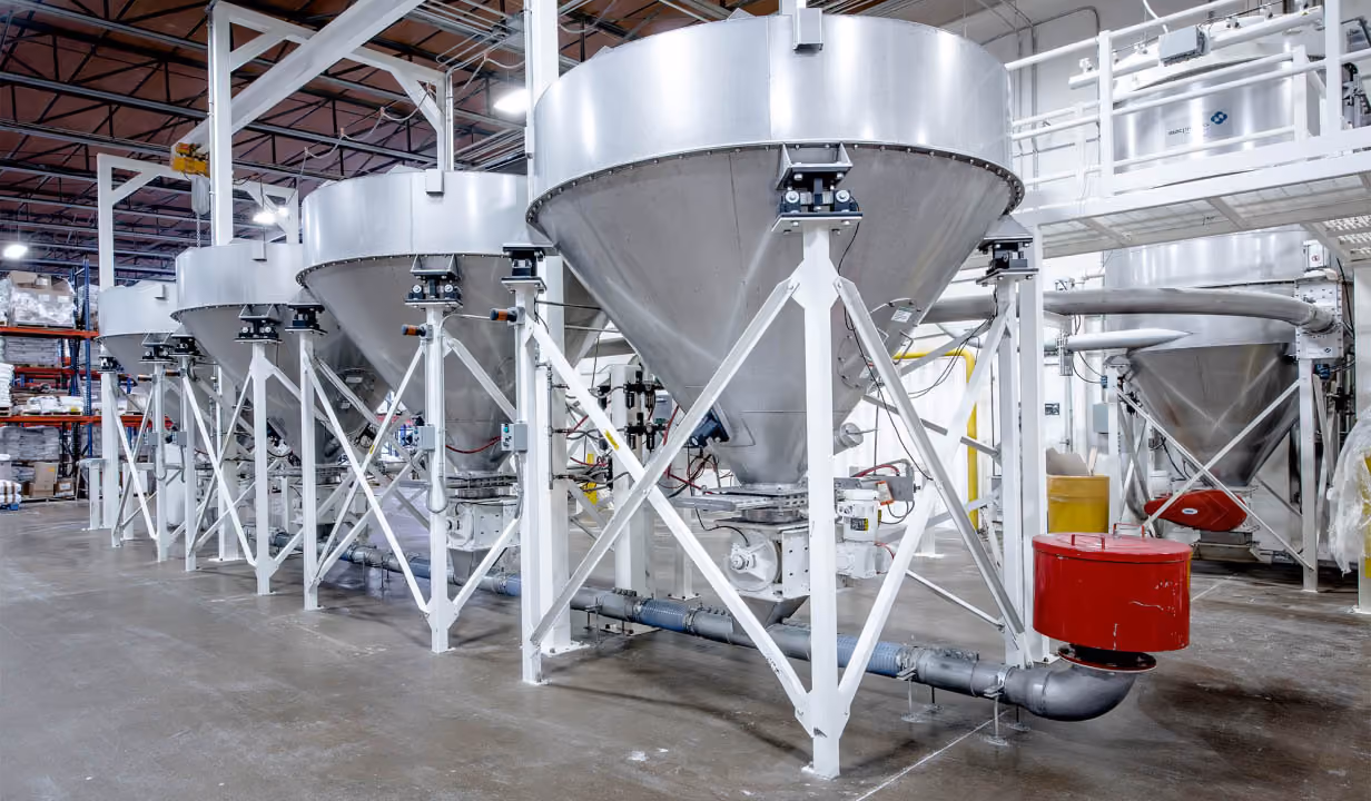 Industrial stainless steel funnel-shaped, custom bakery mix tanks aligned in a warehouse with white support frames.