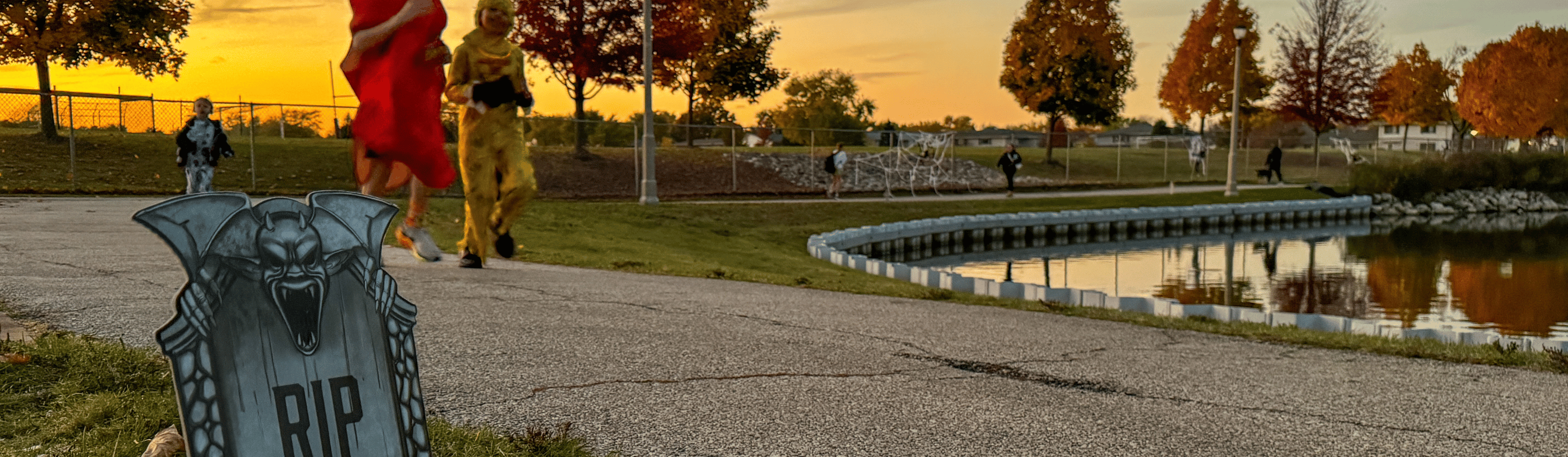 Runners passing a "RIP" tombstone decoration on a pond path at Volkening Heritage Farm at sunset.