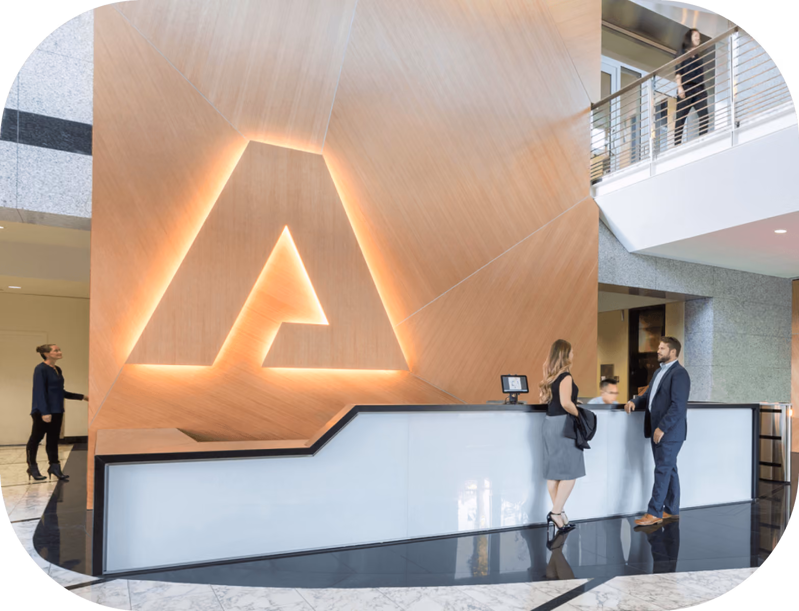 Modern office reception area with a large illuminated 'A' on wood paneling and three people interacting at the desk.
