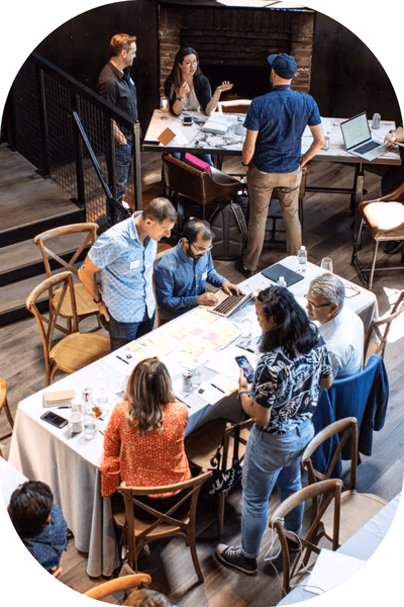 Several people collaborating and discussing around tables with laptops, papers, and smartphones in a modern indoor workspace.
