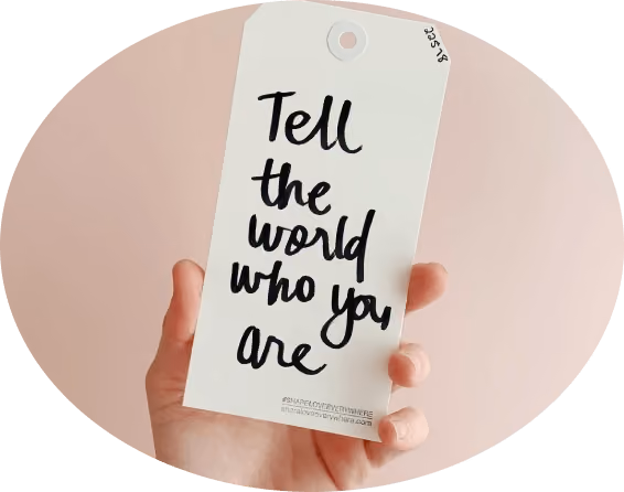 Hand holding a white tag with the handwritten message 'Tell the world who you are' against a pale pink background.