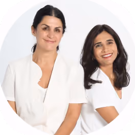 Two women smiling, one with dark hair tied back wearing a white cardigan, and the other with long dark hair wearing a white shirt.